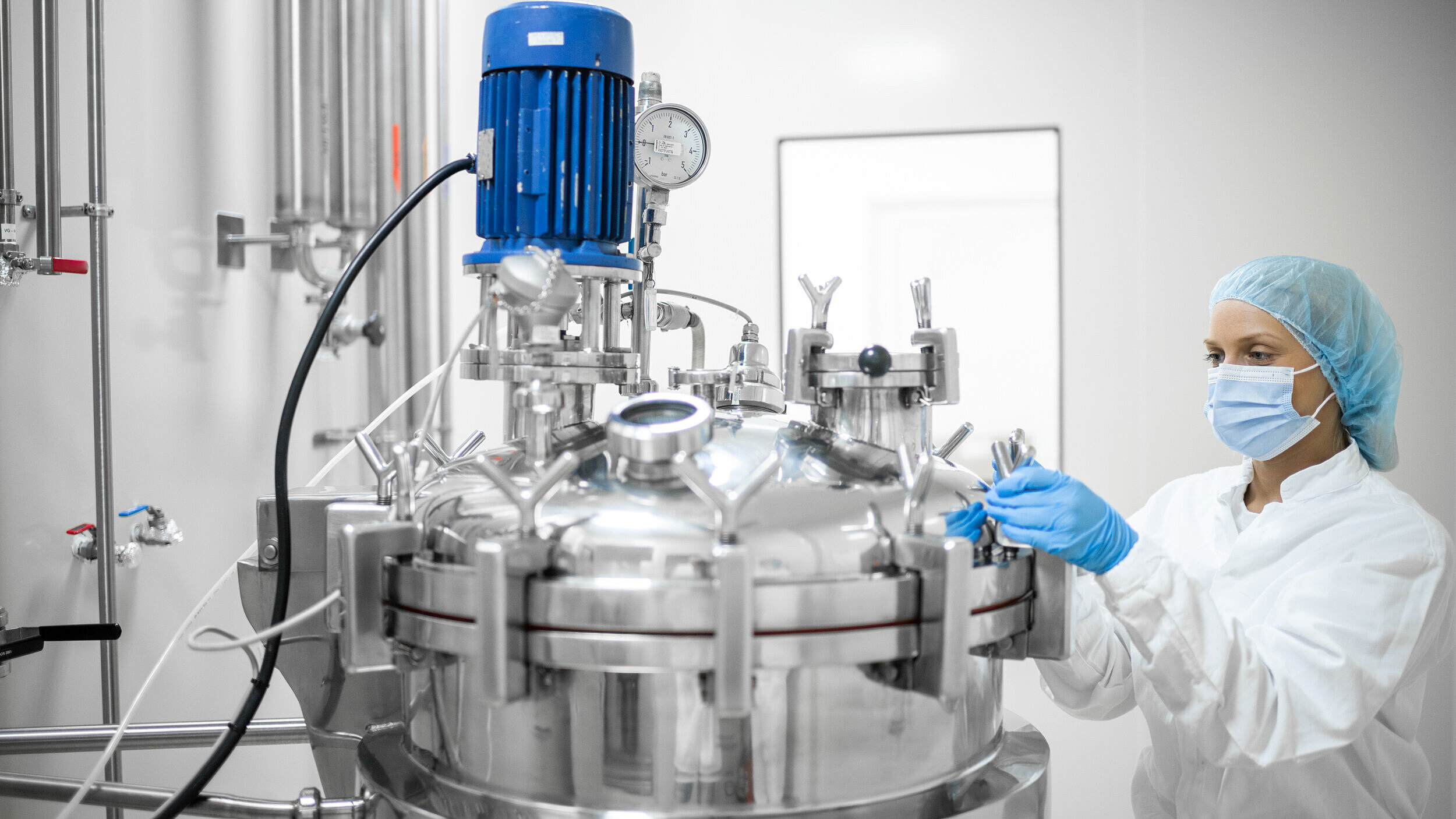 „Mitarbeiterin in Schutzkleidung arbeitet an einem modernen Edelstahltank in einem sterilen Labor oder Pharmaunternehmen – Biotechnologie, pharmazeutische Produktion, Reinraumtechnik